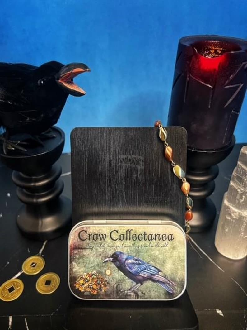 May include: A black crow figurine with an open beak sits on a black pedestal. A tin box with the text "Crow Collectanea" and a crow illustration is in front of a dark wooden stand. A beaded necklace and a red candle are also visible.