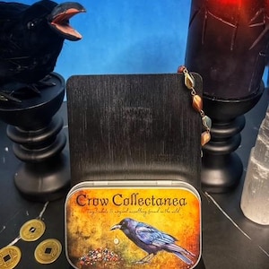 May include: A decorative tin with the text "Crow Collectanea" and a crow illustration. The tin is displayed on a black wooden stand, with a black crow figurine and a lit candle in the background.