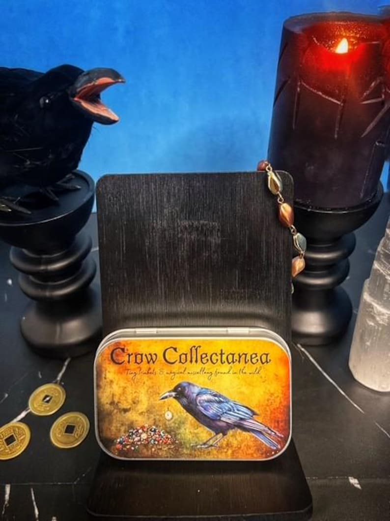 May include: A decorative tin with the text "Crow Collectanea" and a crow illustration. The tin is displayed on a black wooden stand, with a black crow figurine and a lit candle in the background.