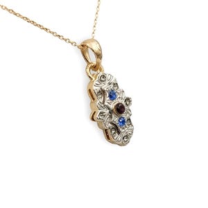 Può includere: Una collana color oro con un ciondolo con un design floreale. Il ciondolo ha un rubino centrale e due zaffiri blu, circondati da piccole pietre preziose trasparenti. La collana è su sfondo bianco.