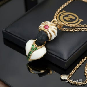 Puede incluir: Un collar de cadena dorada con un colgante en forma de busto que lleva un turbante blanco con una piedra roja central. La figura tiene un rostro negro y una prenda blanca adornada con una fila vertical de piedras verdes, colgada de una cadena.