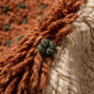 May include: Close-up of a textured, hand-knitted textile. The design features a combination of rust-colored and cream-colored yarn, with small, dark green, flower-shaped embellishments. The fringe adds a rustic touch to the knitted item.