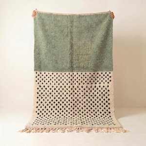 Peut inclure: Un tapis rectangulaire au design bicolore. La moitié supérieure est d'un vert sauge uni avec un poil long et texturé. La moitié inférieure présente une base blanc cassé avec un motif répétitif de petits losanges noirs et une frange.