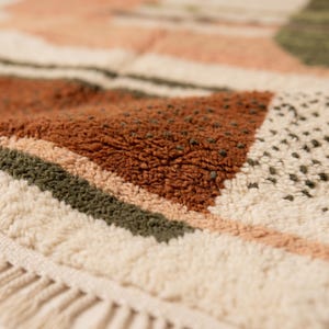 May include: Close-up of a textured rug with a geometric pattern. The rug features a combination of colors, including cream, rust, olive green, and peach. The design includes stripes and triangular shapes, with a fringed edge.