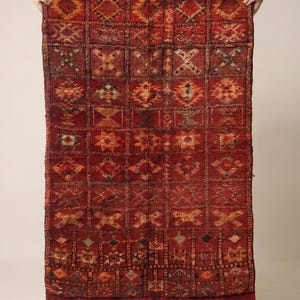 May include: A rectangular, hand-woven rug with a deep red background and a repeating geometric pattern in shades of orange, brown, and black. The rug has a fringe along the bottom edge and is held up by unseen hands. The rug is likely made of wool.
