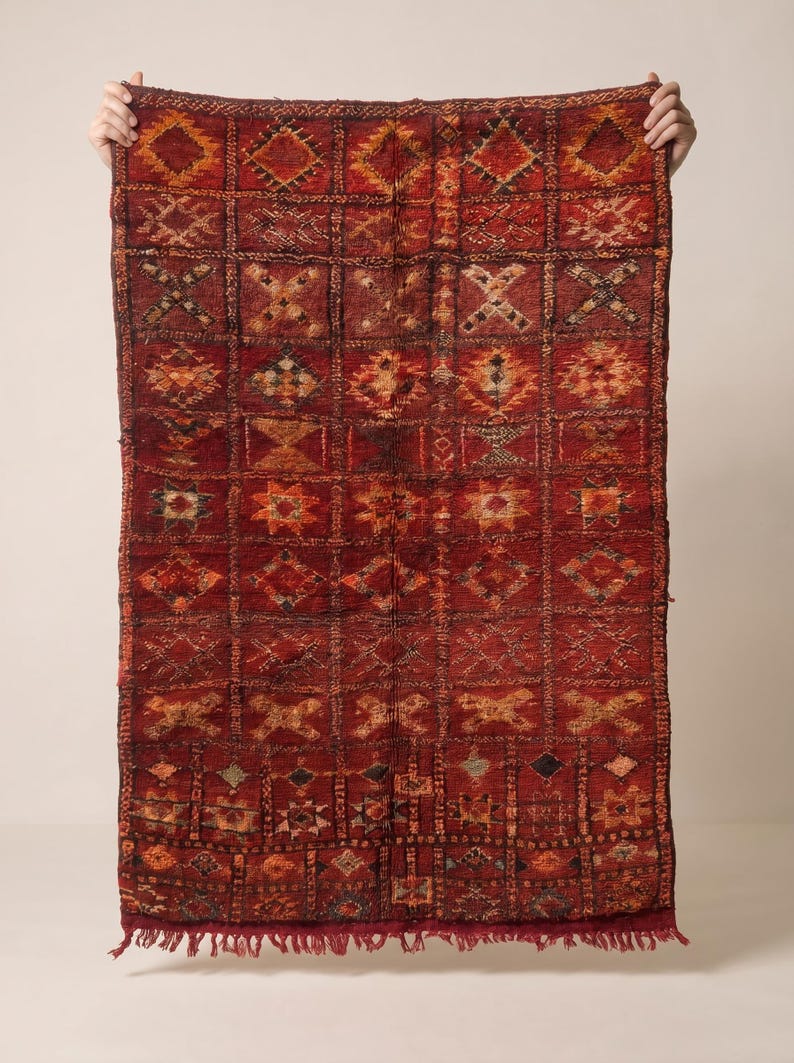 May include: A rectangular, hand-woven rug with a deep red background and a repeating geometric pattern in shades of orange, brown, and black. The rug has a fringe along the bottom edge and is held up by unseen hands. The rug is likely made of wool.