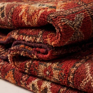 May include: Close-up of a folded, textured rug with a geometric pattern. The rug features a colour palette of red, orange, and brown hues. The fabric appears thick and plush, with visible woven details.