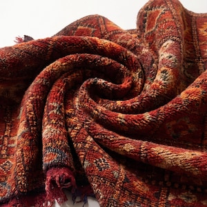 May include: A rolled-up rug with a textured surface, displaying a vibrant mix of reds, oranges, and browns. The rug has a geometric design and fringed edges. The image emphasizes the intricate details and the soft, plush feel of the rug.