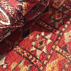 May include: Close-up of a folded, textured rug with a vibrant red base and intricate geometric patterns in black, orange, and cream. The rug's design features repeating shapes and a plush, woven texture.