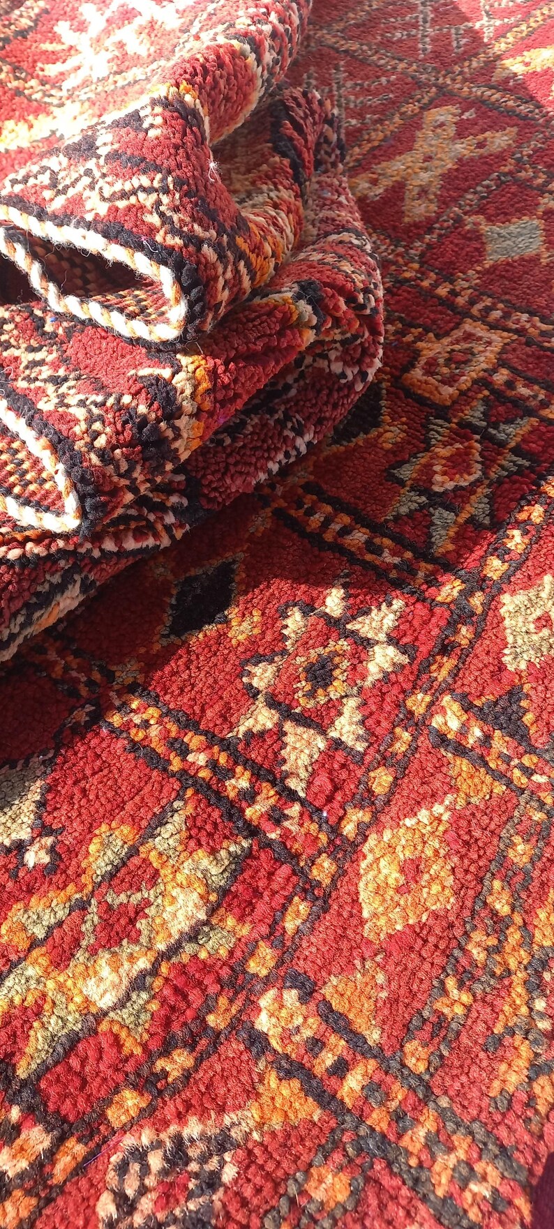 May include: Close-up of a folded, textured rug with a vibrant red base and intricate geometric patterns in black, orange, and cream. The rug's design features repeating shapes and a plush, woven texture.