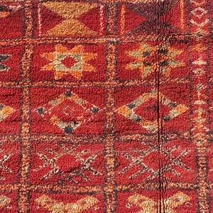 May include: Close-up of a red and orange patterned rug. The rug features a repeating geometric design with squares and rectangles. The pattern includes diamond, cross, and star shapes in shades of orange, black, and white.