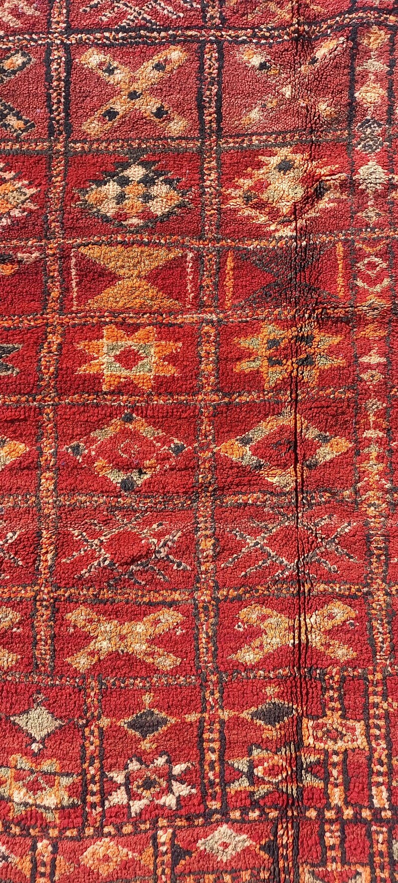 May include: Close-up of a red and orange patterned rug. The rug features a repeating geometric design with squares and rectangles. The pattern includes diamond, cross, and star shapes in shades of orange, black, and white.