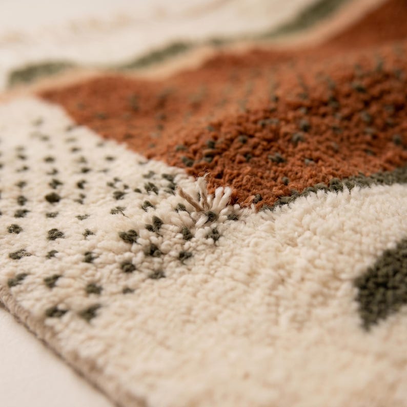May include: Close-up of a textured rug with a geometric pattern. The rug features a combination of cream, rust, and olive green colors. The design includes dotted and striped sections, creating a layered effect. The rug's fibers are visible, adding to its tactile appearance.