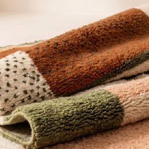 May include: Close-up of several folded, textured rugs. The rugs feature a patchwork design with sections of brown, green, and cream colors. The rugs have a soft, plush appearance, suggesting a comfortable texture.
