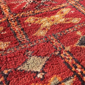 May include: Close-up of a vibrant, textured rug with a geometric pattern. The rug features a rich red base colour with accents of orange, black, and green. The design is intricate and detailed, showcasing a traditional style.