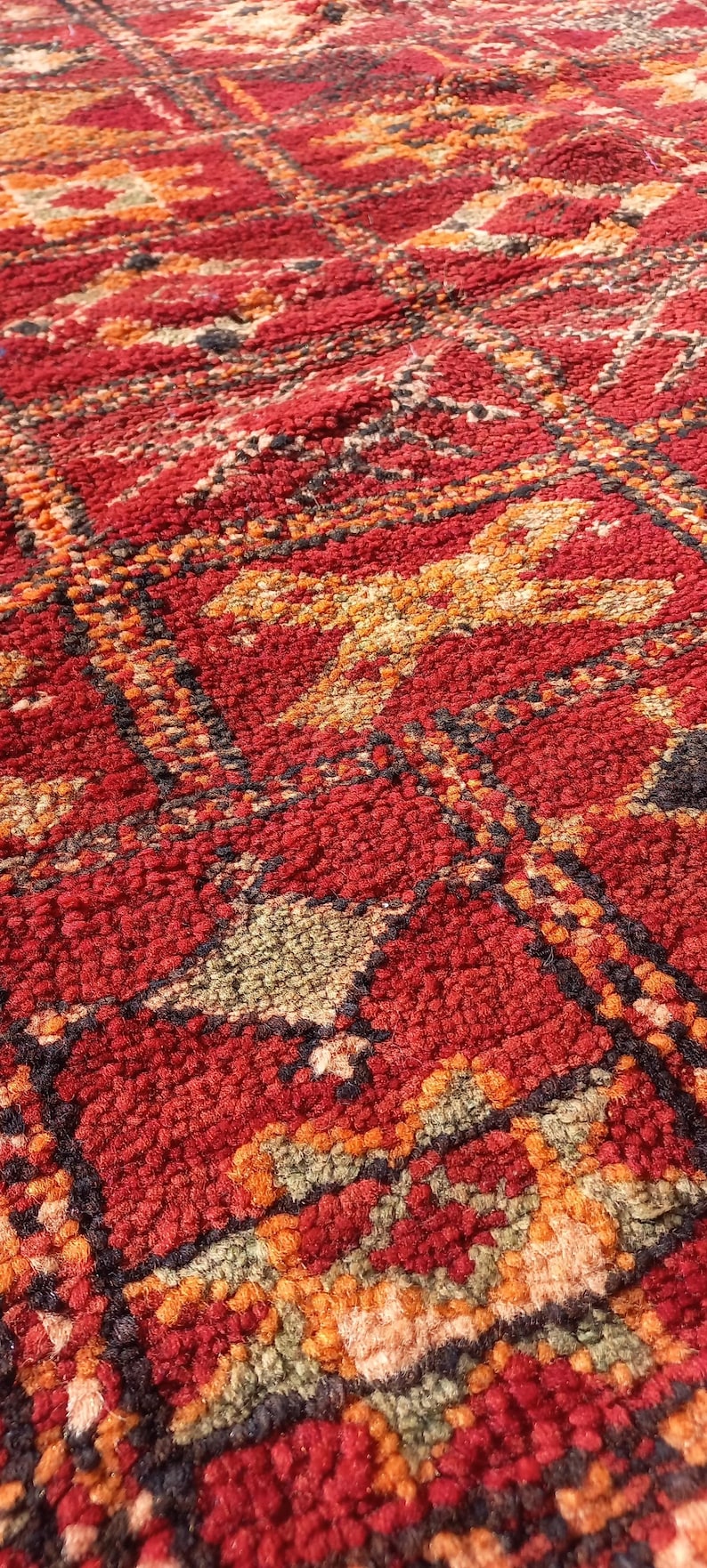 May include: Close-up of a vibrant, textured rug with a geometric pattern. The rug features a rich red base colour with accents of orange, black, and green. The design is intricate and detailed, showcasing a traditional style.