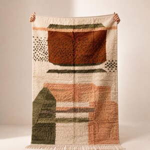 May include: A rectangular rug with an abstract design. The rug features a cream-colored background with geometric shapes in shades of brown, green, and peach. The rug has a fringed edge and is held up by two hands.