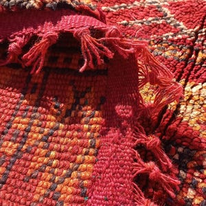 May include: Close-up of a vibrant, hand-woven rug with a rich, textured surface. The rug features a complex geometric pattern in shades of red, orange, and brown, with fringed edges. The design showcases traditional craftsmanship.