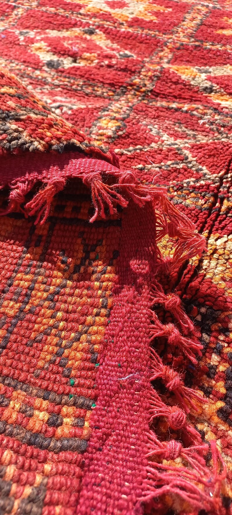 May include: Close-up of a vibrant, hand-woven rug with a rich, textured surface. The rug features a complex geometric pattern in shades of red, orange, and brown, with fringed edges. The design showcases traditional craftsmanship.
