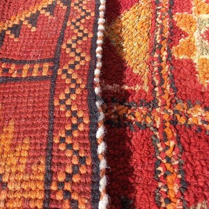 May include: Close-up of a section of a woven rug with a geometric pattern. The rug features a colour palette of red, orange, black, and white. The design includes repeating shapes and lines.