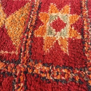 May include: Close-up of a textured rug with a repeating geometric pattern. The rug has a red background with orange, yellow, and black accents. The design includes star-like and square motifs.