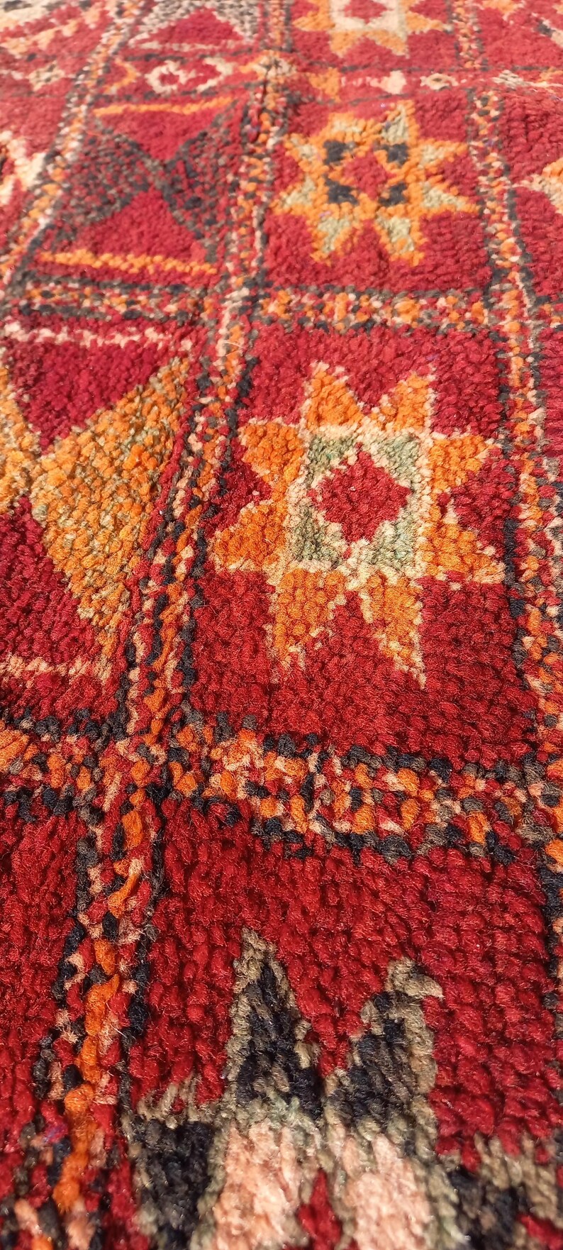 May include: Close-up of a textured rug with a repeating geometric pattern. The rug has a red background with orange, yellow, and black accents. The design includes star-like and square motifs.