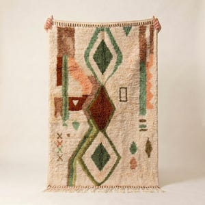 May include: Cream-coloured rug with a geometric design. The rug features a central diamond pattern in shades of green and brown, with accents of peach and brown. The rug is rectangular and has a fringed edge.