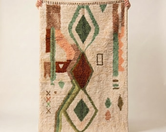 May include: Cream-colored rug with a geometric design. The rug features a central diamond pattern in shades of green and brown, with accents of peach and brown. The rug is rectangular and has a fringed edge.