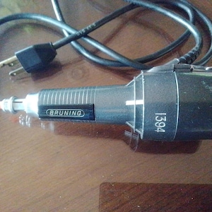 May include: A black and silver Bruning 1394 tool with a power cord and plug. The tool has a cylindrical body with the brand name "Bruning" and the number "1394" printed on it. The tool has a silver tip with an orange end.
