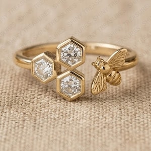 Op de afbeelding: Een open gouden ring met drie zeshoekige zettingen, elk met een heldere ronde edelsteen, naast een kleine gouden bijenfiguur. De ring heeft een gepolijste afwerking en een open ontwerp met de bij en de zeshoeken aan de uiteinden.
