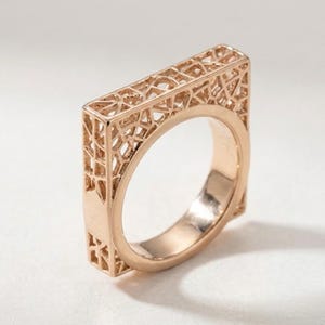 May include: A rose gold ring featuring a square-shaped top section with an intricate, openwork lattice pattern. The band is smooth and circular on the interior, while the exterior top portion displays a geometric, web-like structure.