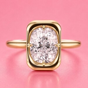 May include: A gold ring featuring a rectangular bezel setting with rounded corners that secures a large, clear, faceted cushion-cut gemstone. The metal band is smooth and polished, showcasing a high-shine finish that reflects light across the surface.