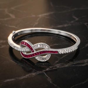 Op de afbeelding: Een zilverkleurige scharnierende armband met een centraal knoopontwerp in de vorm van een oneindigheidsteken. De knoop is versierd met in kanaal gezette, baguette-geslepen rode robijnen en witte stenen, met ronde witte stenen op de band.