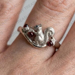 Könnte beinhalten: Ein silberfarbener Ring mit einer skulpturalen Eichhörnchenfigur, die eine dunkelrote Eichel hält. Das Eichhörnchen hat einen kleinen blauen Stein als Auge, und zwei weitere dunkelrote, eichelförmige Steine sind in die texturierte Ringschiene eingesetzt.