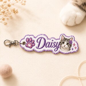 May include: A custom embroidered keychain featuring a purple paw print, the name Daisy in purple script, and a portrait of a tabby cat with a small purple flower on its head. The keychain is finished with a silver metal lobster clasp attachment.