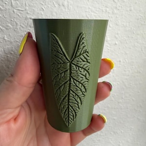 May include: A small, olive green cylindrical cup featuring a raised, textured relief of a single heart-shaped leaf on its front surface. The cup has a smooth, matte finish and a tapered shape that narrows slightly toward the flat base.