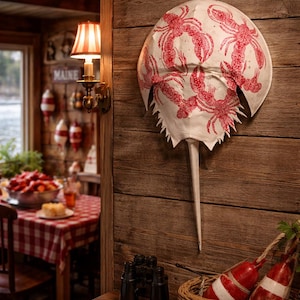 以下が含まれることがあります： 赤いロブスターのイラストが描かれた、大きな白いカブトガニ型の壁掛け。木製の壁に取り付けられています。小さなテーブルには双眼鏡と、赤と白のブイが入ったバスケットが飾られています。