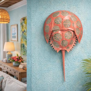 May include: Coral-colored horseshoe crab wall art with green circular patterns. The decorative piece is mounted on a textured, light blue wall. A framed picture of a beach scene hangs nearby, with a glimpse of a coastal interior.