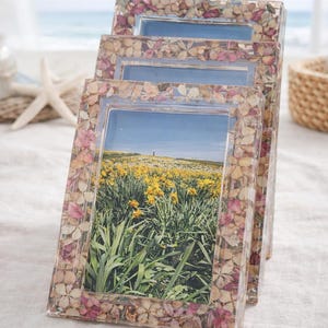 Op de afbeelding: Een set van drie rechthoekige fotolijsten. De lijsten zijn versierd met gedroogde bloemen in roze, beige en wit. Elke lijst bevat een foto van een veld met gele bloemen onder een blauwe lucht.