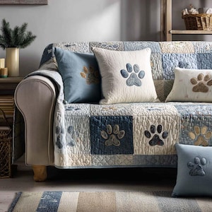 May include: A sofa with a patchwork quilt and several throw pillows. The quilt and pillows feature paw print designs in shades of blue, beige, and brown. The sofa has a light beige frame and is placed in a cozy living room setting.