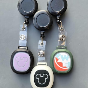 Puede incluir: Tres portatarjetas retráctiles con carretes negros y correas de plástico transparente. Uno tiene una cara lavanda con una silueta blanca de Mickey Mouse, otro tiene una cara blanca con una silueta negra de Mickey Mouse, y el tercero tiene una cara verde con un gráfico.