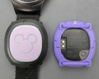 Puede incluir: Un reloj inteligente negro con correa negra y un inserto circular lavanda con la silueta blanca de la cabeza de Mickey Mouse, junto a una funda protectora lavanda que muestra la parte trasera negra de un dispositivo con pines de carga dorados.