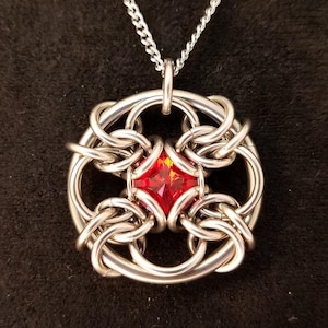 May include: A silver chain necklace with a round pendant featuring a red gemstone set in a silver, intricate, woven design.