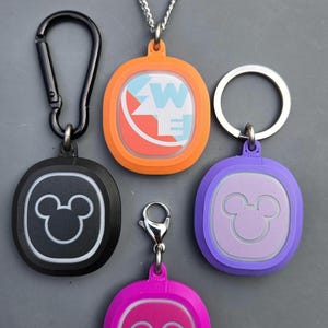 May include: A selection of keychains and pendants in various colours. The pendants are in orange, black, purple, and pink, each with a different design. One has a carabiner clip, one has a chain, and one has a keyring.