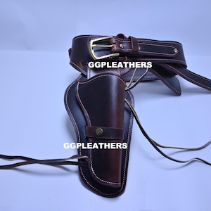 Może przedstawiać: Brązowy skórzany zestaw kabury i paska do pistoletu ze złotą klamrą. Kabura jest przeznaczona do przechowywania pojedynczej broni palnej i jest przymocowana do szerokiego paska. Tekst "GGLEATHERS" jest widoczny na kaburze.