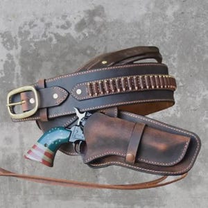 Puede incluir: Un cinturón de cuero marrón con una funda y un revólver. El revólver tiene un mango azul, rojo y blanco. El cinturón tiene una hebilla dorada y una fila de balas.