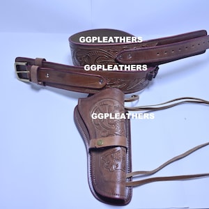 May include: A brown leather gun holster set, including a belt and a wide band, with embossed floral patterns. The set features the text "GGLEATHERS". The belt has a gold-coloured buckle. The holster has a strap with a button closure.