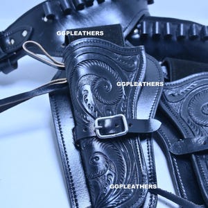 Può includere: Fondina per pistola in pelle nera con design floreale in rilievo. La fondina è dotata di chiusura con fibbia ed è contrassegnata con la scritta "GGLEATHERS". L'immagine mostra un primo piano della fondina e della cintura.