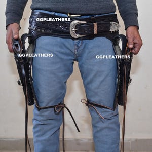 Peut inclure: Holster de pistolet en cuir noir avec une ceinture décorative et une boucle argentée. L'étui est attaché aux jambes avec des lanières de cuir. Le texte "GGLEATHERS" est visible sur l'étui et la ceinture.