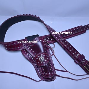 Może przedstawiać: Bordowy skórzany zestaw kabury i paska do pistoletu. Zestaw zawiera kaburę, pasek i pasek na ramię, wszystkie ozdobione srebrnymi ćwiekami. Pasek ma zapięcie na klamrę. Tekst "GGLEATHERS" jest widoczny na pasku.